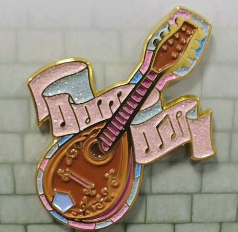 Soft enamel pin in the shape of a bard's mandolin, with scroll detail on the body and pink frets along the neck. It's surrounded by a thin stained glass background in the shades of pink, blue & white. There is also a pink scroll with musical notes flowing behind the neck of the mandolin. The stained glass area and music scroll  have pink, blue and white glitter.