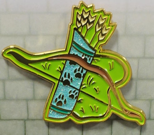 Soft enamel pin depicting the weapon of a Ranger. It has a glittery teal quiver decorated with bear paw prints, and holding a few arrows. There is also a brown bow laying on the quiver, and both are displayed against a patch of green grass.