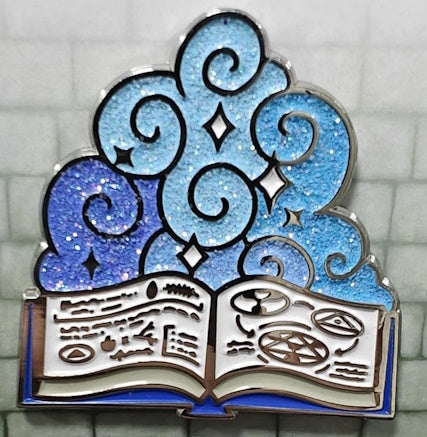Soft enamel pin showing a Wizard's open spell book, with 3 shades of glittery blue magic swirling above it.