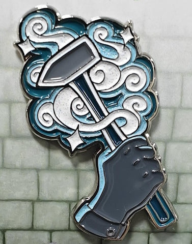 Soft enamel pin depicting an Artificer's gray hand holding a gray and white hammer with a teal handle, with swirls of white glitter, and light & dark teal swirling behind the hammer.